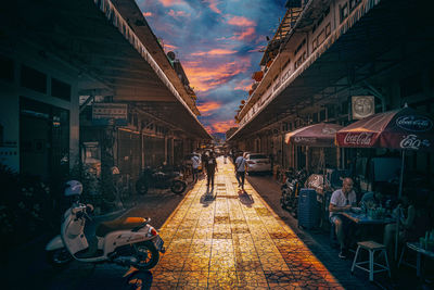 People sitting on sidewalk by buildings against sky during sunset