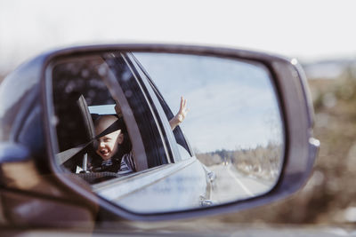 Car mirror shot of boy in back seat with hand out window on sunny day