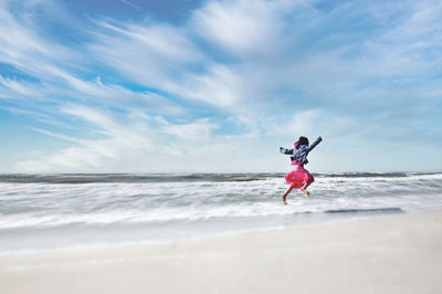 Full length of girl jumping while running at beach against sky