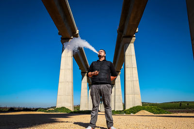 Low angle view of man smoking electronic cigarette while standing on sand against blue sky