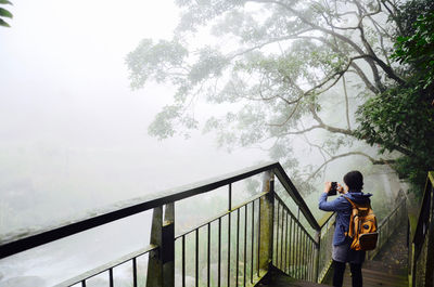 Rear view of woman photographing with mobile phone while standing on steps during foggy weather