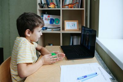 8 years old boy sit by desk with laptop and do exercise with massage ball. side view. smiling woman