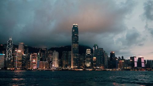 Illuminated city at waterfront against cloudy sky