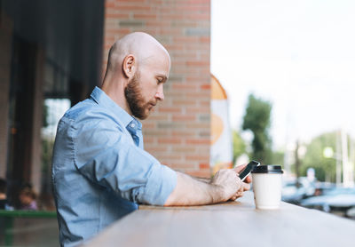 Adult attractive man forty years with beard in blue shirt businessman using mobile phone at cafe