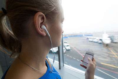 Young woman looking through window while listening music at airport
