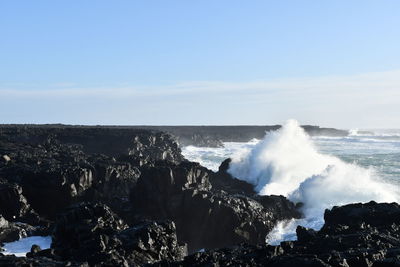 Waves splashing on rocks at shore against sky