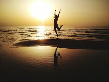 Silhouette woman with arms raised jumping at beach against sky during sunset