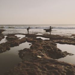 Full length side view of men standing on rock formation at beach against sky