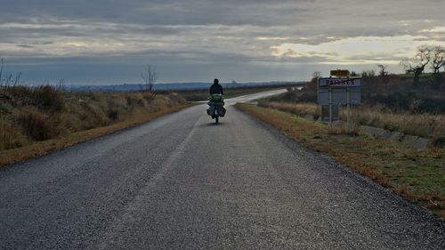 Rear view of man riding bicycle on road against cloudy sky during sunset