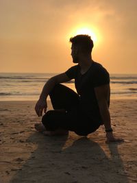 Man sitting on beach against sky during sunset