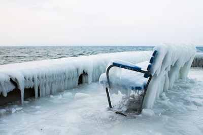 Frozen bench by sea against sky