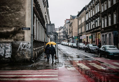 Rear view of man and woman walking on wet sidewalk by buildings