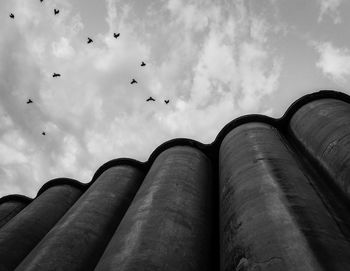 Low angle view of birds flying over silos against cloudy sky