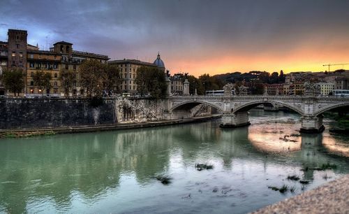Bridge over river by buildings against sky at sunset