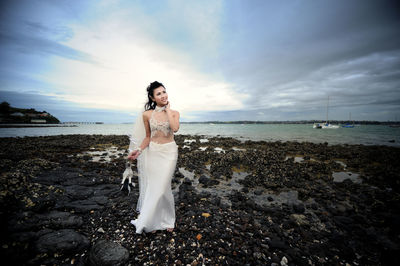 Full length of smiling bride holding high heels while standing at beach
