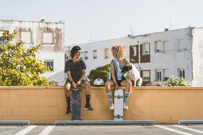 Friends with skateboard and dog talking while sitting on retaining wall in city