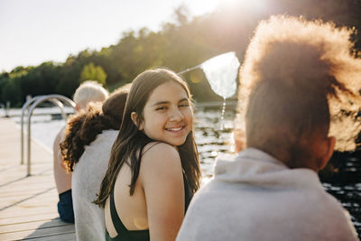 Portrait of smiling girl sitting with friends on jetty by lake