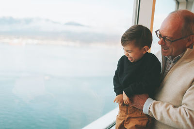Cheerful man holding grandson looking through window against sea