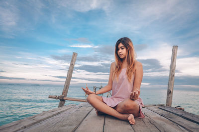 Mid adult woman meditating while sitting on pier over sea against sky