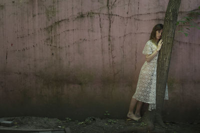 Portrait of young woman standing by tree against wall