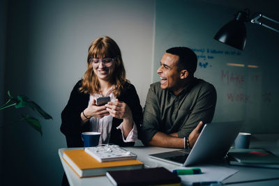 Cheerful multi-ethnic business colleagues sitting at desk with technologies at creative office