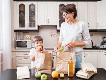 Woman and toddler in kitchen. kid bites apple. grocery delivery in paper bags. mother and son.