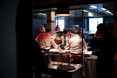 Young cooks men in aprons carefully serving meal in white ceramic dishes on restaurant kitchen