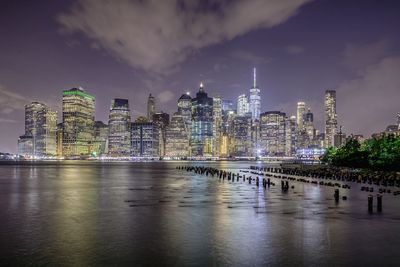Illuminated modern buildings by river against sky at night