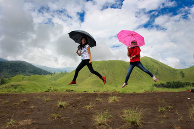 Portrait of happy friends with umbrellas jumping on field against cloudy sky