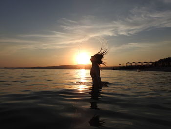 Woman tossing hair while standing in sea against sky during sunset
