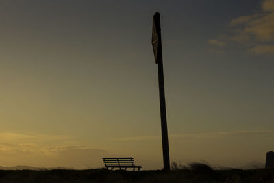 Silhouette pole and empty bench on field against sky at sunset