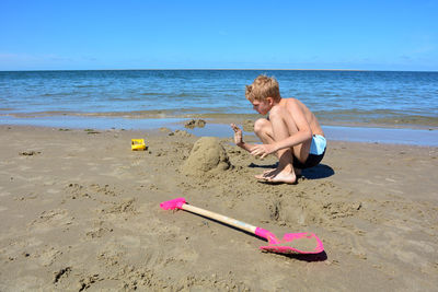 A little blond boy with swimming trunks builds a sand castle in the sand on the beach 