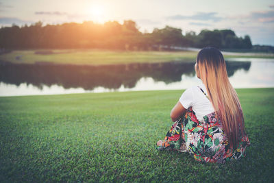 Rear view of woman looking at lake while sitting on field during sunset