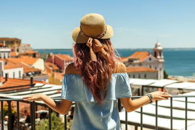 Rear view of young woman looking at buildings and sea while standing by railing in balcony against clear sky