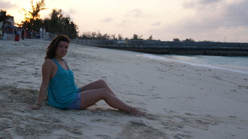 Young woman sitting at beach against sky during sunset