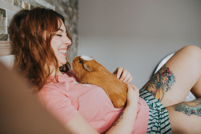 Side view of young woman in pajamas playing with guinea pig and relaxing on bed in bedroom