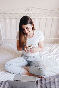 Young woman using phone while sitting on bed at home