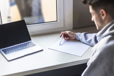 High angle view of man writing over paper by laptop on table