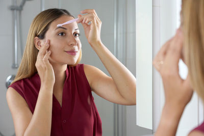 Hispanic woman shaving her eyebrows with razor in the mirror at home