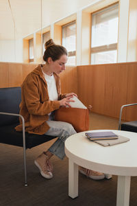 Full body young female student in casual clothes with hair bun taking stationery from bag while sitting on chair near table and preparing for studies in university library