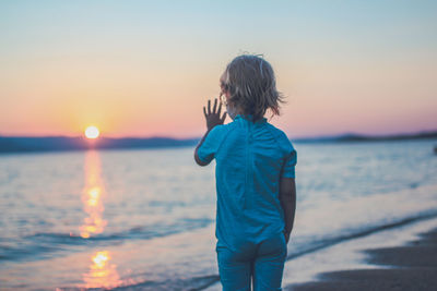 Rear view of boy standing on beach against sky during sunset