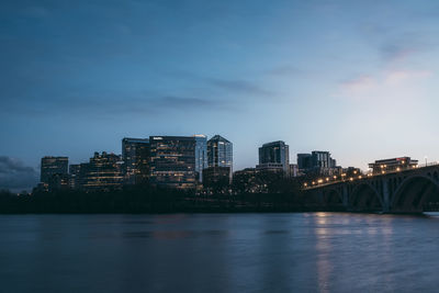 Illuminated buildings by river against sky at dusk