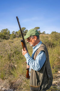 Side view of male hunter in special hunting vest and green cap holding rifle and looking away among tall grass