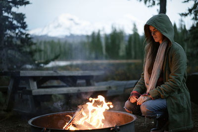 Woman crouched by campfire with hood up