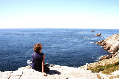 Rear view of woman sitting on cliff by sea against clear sky