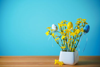 A bouquet of spring yellow flowers on blue background with easter eggs with pattern in a white vase.