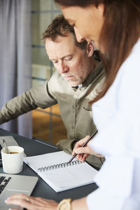 Mature creative colleagues writing in dairy while sitting with laptop at conference table in board room