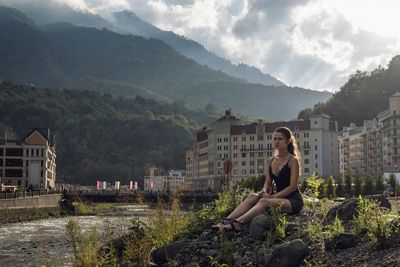 Young woman sitting on built structure against trees