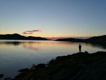 Silhouette man standing on rock by lake against sky during sunset