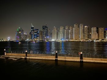 Illuminated buildings by sea against sky at night